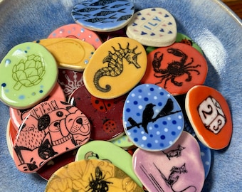 May include: A collection of colourful ceramic tiles with various designs, including a seahorse, a crab, a ladybug, a bird, a bee, a mouse, a dog, an artichoke, and the number 23. The tiles are in a blue bowl.