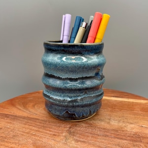 Könnte beinhalten: Ein handgefertigter, blauer Keramik-Stifthalter mit einem strukturierten, geriffelten Design. Der Stifthalter enthält mehrere farbige Stifte in Lila, Blau, Grau, Rot und Gelb. Der Stifthalter steht auf einer Holzoberfläche.