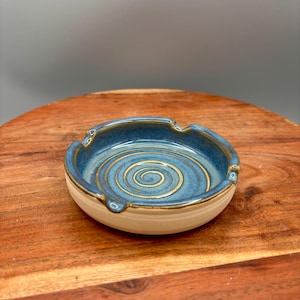 以下が含まれることがあります： 青色の内側とクリーム色の外側を持つ手作りのセラミック灰皿。内側は、より明るい青と金の色のスパイラルデザインが特徴です。灰皿には、タバコを置くための3つの切り込みがあります。