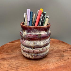 May include: A handmade ceramic pen holder with a textured, ridged design in shades of burgundy, cream, and brown. The holder is filled with various pens and markers in different colors. The pen holder sits on a wooden surface.