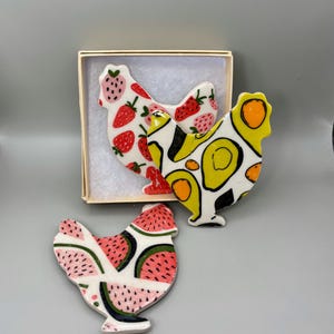 May include: Three ceramic chicken-shaped decorations. One chicken is decorated with strawberries, another with avocados, and the third with watermelon slices. The chickens are white with colorful fruit designs and black accents. One chicken is in a small box.