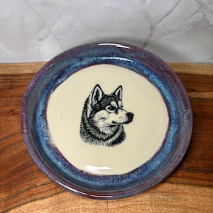 Könnte beinhalten: Ein runder Keramikteller mit cremefarbenem Zentrum, der eine schwarz-weiße Illustration des Kopfes eines Husky-Hundes zeigt. Der Rand des Tellers hat einen Farbverlauf aus Blau- und Lilatönen.