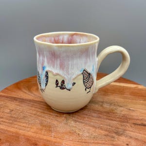 May include: Cream-colored ceramic mug with a light pink and white glaze on the rim. The mug features hand-drawn bird illustrations in black ink with blue and red accents. The handle is a matching cream color.