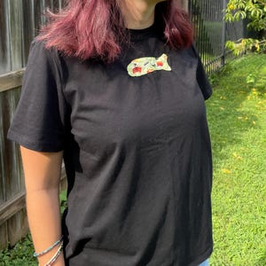 May include: Black t-shirt with a small, colorful graphic on the chest. The graphic appears to be a stylized food item. The shirt is short-sleeved and made of a dark fabric. The background includes a wooden fence and green grass.