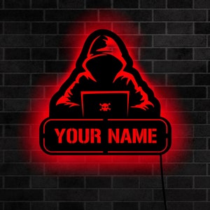 May include: Red neon sign featuring a silhouette of a hooded figure seated at a laptop with a skull and crossbones on the screen. The sign reads "YOUR NAME" at the bottom.