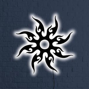 Può includere: Opera d'arte da parete a forma di sole nero stilizzato con un contorno bianco luminoso. Il design presenta forme a fiamma che si irradiano da un cerchio centrale. L'opera è montata su un muro di mattoni blu scuro, con un cavo di alimentazione visibile.