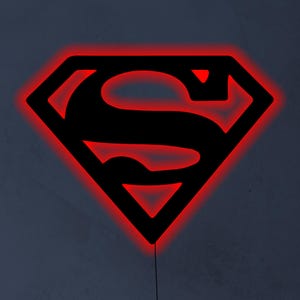 May include: Black Superman logo with a red neon outline. The iconic "S" shield is illuminated with a vibrant red glow, creating a striking contrast against the black emblem. A cord extends from the bottom.