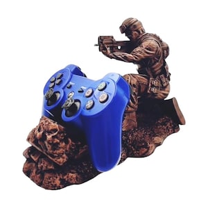 May include: A blue video game controller is held by a brown figurine of a soldier holding a gun. The soldier and controller are on a brown rock base.