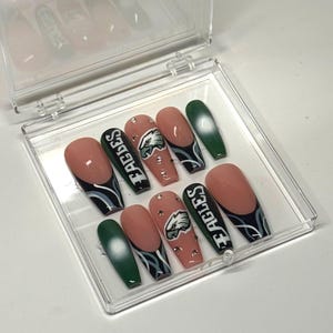 May include: A set of artificial nails in a clear plastic case. The nails feature a variety of designs, including a pink base with a white and navy blue French tip, a green base, and nails with the text "EAGLES" and a team logo.