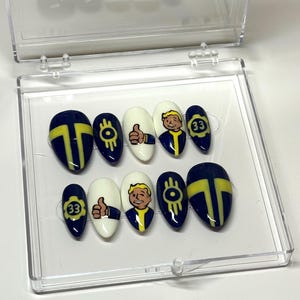 May include: A set of press-on nails in a clear plastic case. The nails feature navy blue and yellow designs, including a thumbs-up, the number 33, and a cartoon character. Some nails are white.