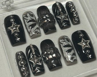 May include: A set of ten press-on nails in a clear plastic case. The nails feature a black and white color palette with glossy finishes, metallic silver star charms, raised textured patterns, and small silver spherical studs arranged in vertical lines.