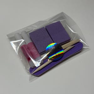 May include: A clear plastic bag containing a nail care kit. The kit includes two purple nail files, two small tubes of pink nail glue, two wooden cuticle sticks, and a holographic sticker with the text "Nail Chic".
