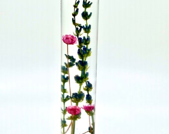 May include: A clear glass vial with a silver cap, filled with dried lavender and small pink flowers. The lavender has purple buds and green stems. The flowers are a vibrant pink. The vial is approximately 15 cm tall.