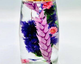 May include: A clear glass bottle with a cork stopper, filled with dried flowers. The arrangement includes purple, pink, and green floral elements. The bottle is cylindrical and sits on a white surface.