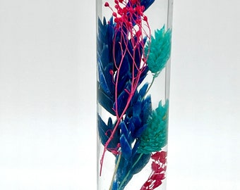 May include: A clear glass bottle with a cork stopper, filled with dried flowers in shades of blue, teal, and red. The flowers are suspended in a clear liquid, creating a decorative display. The bottle is cylindrical and sits on a white surface.