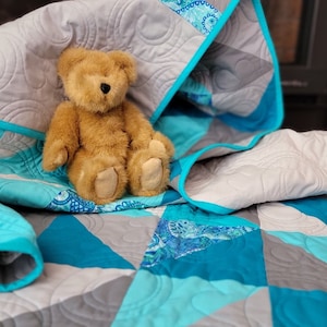 May include: A turquoise and gray patchwork quilt with a teddy bear sitting on top. The quilt has a circular quilting pattern.