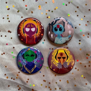 Könnte beinhalten: Vier runde Ansteckbuttons mit farbenfrohen Illustrationen. Jeder Button zeigt ein einzigartiges Charakterdesign mit leuchtenden Farben, darunter Rot, Blau, Grün und Gelb. Die Buttons sind auf einem hellgrünen Hintergrund mit goldfarbenen sternförmigen Konfetti platziert.