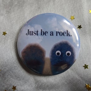 May include: A round button featuring a blue sky with white clouds as the background. The text "Just be a rock." is printed in black. Two fuzzy blue figures with eyes are walking towards each other.
