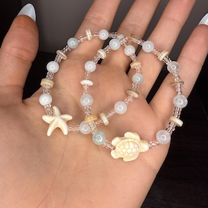May include: Two beaded bracelets with a beach theme. Each bracelet features white, light blue, and clear beads. One bracelet has a starfish charm, and the other has a turtle charm. The bracelets are held in a hand.