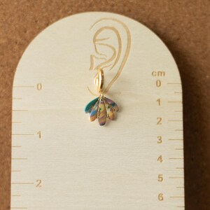 May include: A single gold-colored hoop earring with a colorful, iridescent, petal-shaped charm. The earring is displayed on a wooden measuring tool with centimeter markings. The charm is approximately 1 cm in length.
