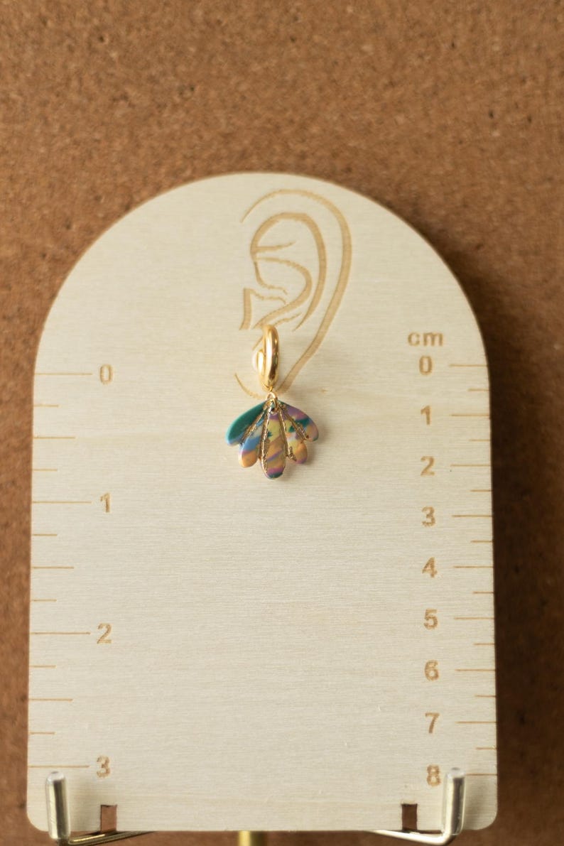 May include: A single gold-colored hoop earring with a colorful, iridescent, petal-shaped charm. The earring is displayed on a wooden measuring tool with centimeter markings. The charm is approximately 1 cm in length.