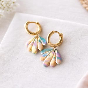 May include: A pair of gold hoop earrings with colorful, fan-shaped pendants. The pendants feature pastel shades of yellow, blue, and pink. The earrings are displayed on a white surface.
