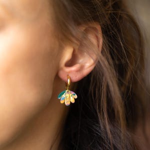 May include: Gold hoop earrings with a colorful, flower-shaped charm. The charm features a mix of pastel colors including yellow, pink, blue, and green. The earrings are a stylish accessory.