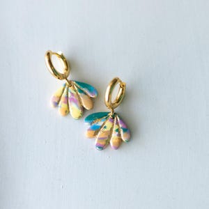 May include: A pair of gold hoop earrings with colorful, fan-shaped pendants. The pendants feature a blend of yellow, pink, blue, and green hues, creating a vibrant, abstract design. The earrings are set against a white background.