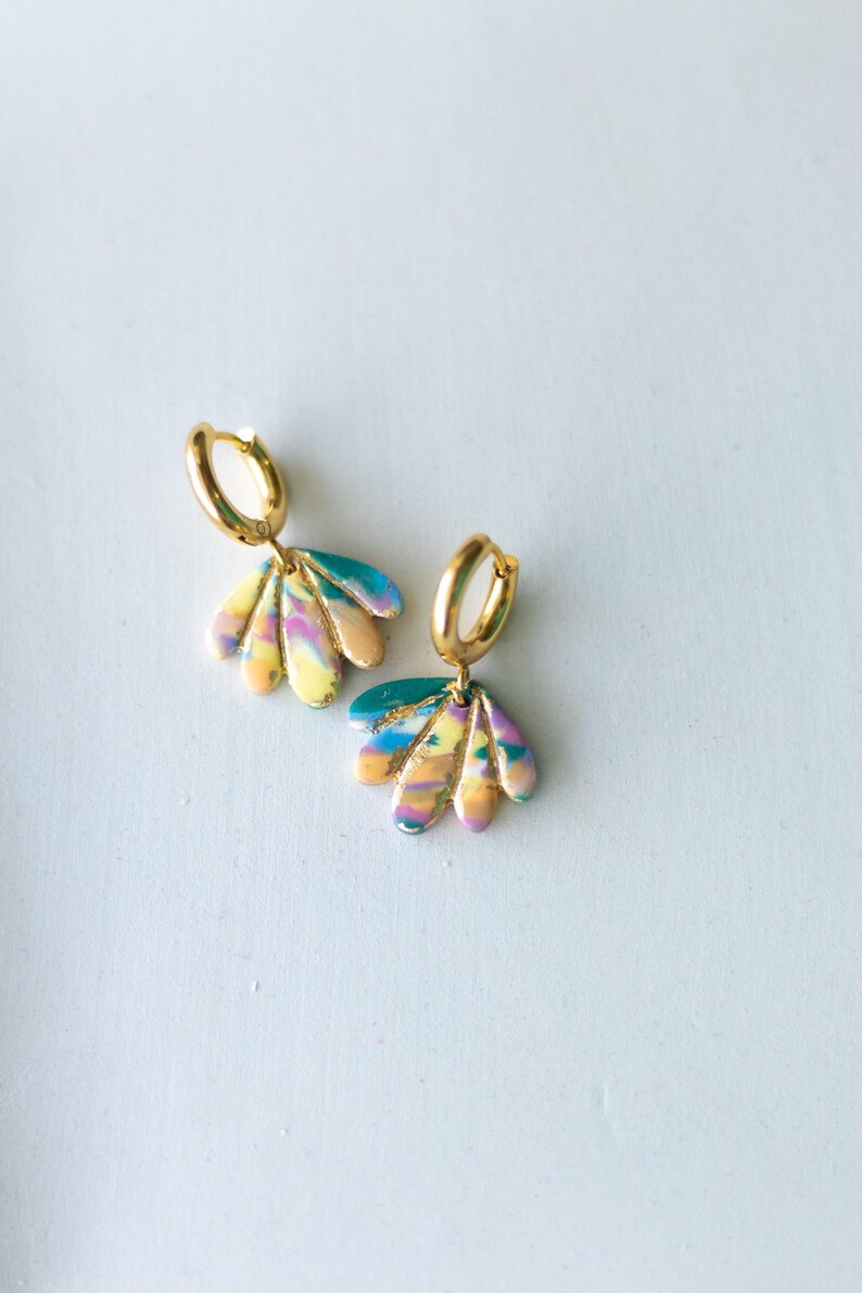 May include: A pair of gold hoop earrings with colorful, fan-shaped pendants. The pendants feature a blend of yellow, pink, blue, and green hues, creating a vibrant, abstract design. The earrings are set against a white background.