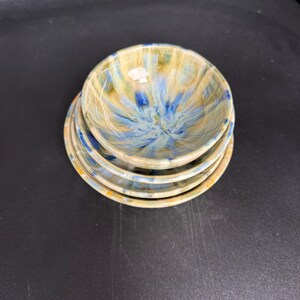 Peut inclure: Une pile de quatre petits bols en céramique. Les bols ont une forme ronde avec un bord légèrement évasé. L'intérieur de chaque bol présente un motif tourbillonnant bleu et jaune, tandis que l'extérieur est de couleur jaune uni.