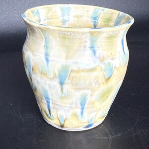 Peut inclure: Un vase en céramique fait à la main avec un bord large et légèrement évasé. Le vase présente un mélange de couleurs, dont le jaune pâle, le bleu clair et le blanc, avec des stries bleues. Le design est organique et texturé.