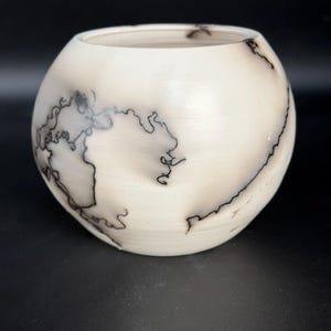 May include: A spherical, cream-coloured ceramic bowl with a distinctive design. The bowl has black, lightning-like patterns across its surface. The interior is smooth and the bowl sits on a black surface. This decorative bowl is ideal for home decor.