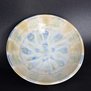Peut inclure: Un bol en céramique fait à la main avec un motif tourbillonnant unique. Le bol présente un mélange de couleurs, notamment bleu clair, vert et beige, créant un effet doux d'aquarelle. Le bol est rond et possède un bord légèrement surélevé.