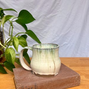 May include: A handmade ceramic mug with a rounded body and a curved handle. The mug features a cream-coloured base glaze with a green drip pattern cascading from the rim. It is resting on a brown hardcover book titled Fifteen Greek Plays.