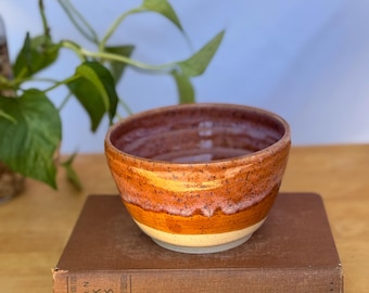 Puede incluir: Un cuenco de cerámica hecho a mano con una cálida paleta de colores naranja, marrón y crema. El cuenco es redondo con un borde ligeramente ensanchado y está sobre un libro antiguo. El interior es de un marrón rojizo oscuro.