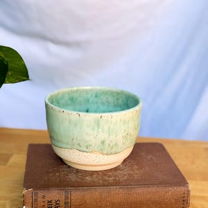 May include: A handmade ceramic bowl with a light green glaze over a cream-colored base. The bowl is sitting on a vintage book titled "Fifteen Greek Plays". The bowl is approximately 3 inches in diameter and 2.5 inches tall.
