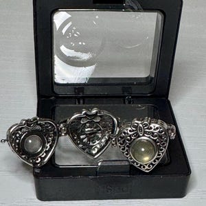 May include: A silver-toned metal ring featuring three heart-shaped components with intricate filigree detailing. The central heart is flanked by two smaller hearts, one of which contains a smooth, round, translucent stone set in the center.