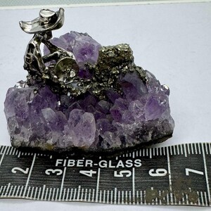 May include: A silver figurine of a skeleton playing a drum, perched atop a cluster of purple amethyst crystals. The amethyst is approximately 3 inches wide. The crystals have a rough, natural texture, and the figurine is detailed.