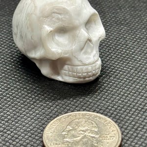 May include: White carved skull made of polished stone. The skull is about the size of a US quarter.