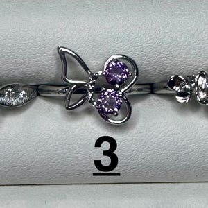 May include: Five silver-toned rings arranged in a row, numbered 1 through 5. Ring 1 features a pear-shaped purple stone with clear accents. Ring 2 has a central oval light blue stone surrounded by clear stones. Ring 3 is a butterfly shape with purple stones. Ring 4 has a cluster of clear stones. Ring 5 features a round clear stone.