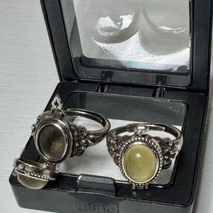 May include: Two silver-toned metal rings with oval-shaped settings inside a black plastic display box. One ring features a hinged locket design that is open, revealing an empty interior, while the other ring is closed with a smooth, pale green stone.