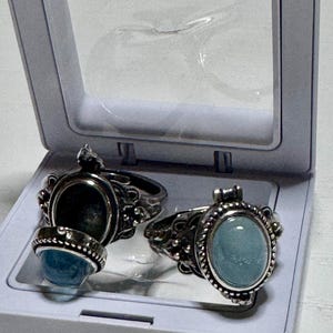 May include: Two silver rings with blue stones. The rings are in a white box with a clear lid. The stones are oval shaped and have a light blue color.