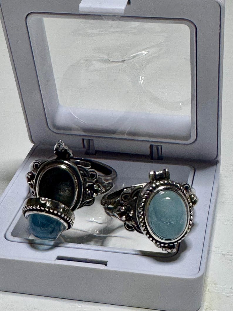 May include: Two silver-toned poison rings featuring oval-shaped light blue cabochon stones. One ring is closed, while the other is open, revealing a hollow compartment beneath the hinged lid. The rings have intricate metalwork and beaded edges.