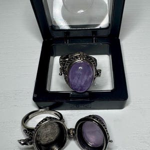 May include: Two silver-toned metal rings with oval purple stones. One ring is closed, displaying the smooth purple stone, while the other is open, revealing a hollow interior compartment. Both rings feature intricate metalwork along the bands.