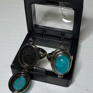 May include: Two silver-toned locket rings featuring oval-shaped, bright turquoise cabochon stones. One ring is closed, displaying the smooth stone, while the other is open, revealing an empty interior compartment. Both rings rest inside a black box.
