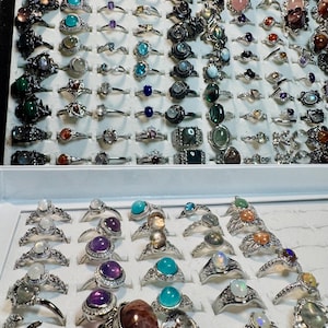 May include: A display of numerous rings with various gemstones, including turquoise, amethyst, and opals. The rings are set in silver and arranged in a display case, showcasing a variety of designs and colors. The collection includes rings with both large and small stones.