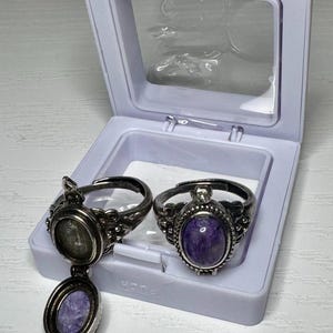May include: Two silver-toned metal rings with oval settings featuring purple stone inlays. One ring is a locket style with an open hinged cover revealing a purple stone, while the other ring is closed, displaying a smooth, polished purple oval stone.