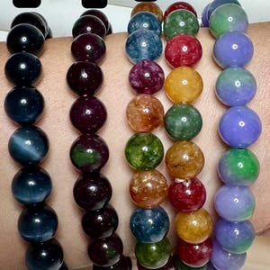 May include: Five beaded bracelets with different colored gemstones. The bracelets are numbered 1 through 5. The beads are round and vary in color, including black, deep red, blue, green, and yellow. The bracelets are displayed on a wrist.
