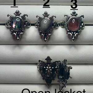 May include: Three silver-toned metal rings with oval-shaped cabochons in iridescent abalone, grey, and reddish-brown stone. Each ring features an ornate, filigree-style setting. Below, one ring is shown in an open locket position, revealing a hollow interior.