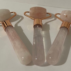 May include: Three rose quartz facial rollers with rose gold accents. The rollers are made of clear quartz with a pink hue. The handles are long and slender.